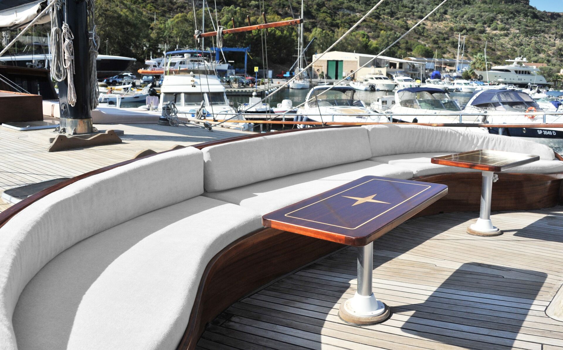 Curved cushioned seating and wooden tables on the deck of a yacht, anchored in a marina with several other boats and lush green hills in the background.