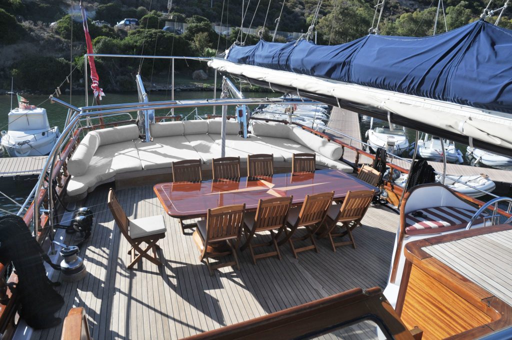 A yacht deck with a long wooden dining table surrounded by 12 chairs, cushioned seating along the edge, and a row of sailboats docked nearby in a marina with lush greenery in the background.