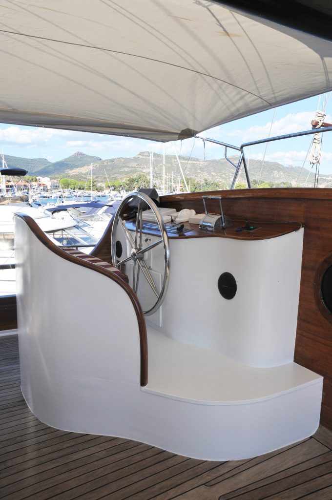 Yacht helm station with a large stainless steel steering wheel, wooden accents, and covered canopy, overlooking a marina with other boats and mountains in the background.