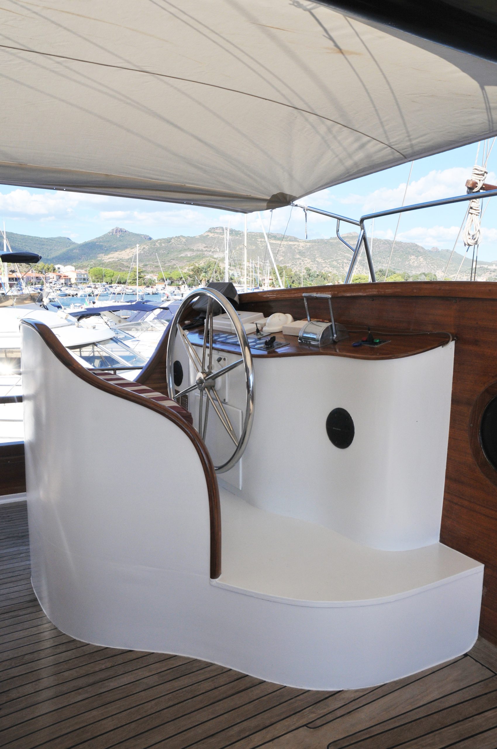 Yacht helm station with a large stainless steel steering wheel, wooden accents, and covered canopy, overlooking a marina with other boats and mountains in the background.
