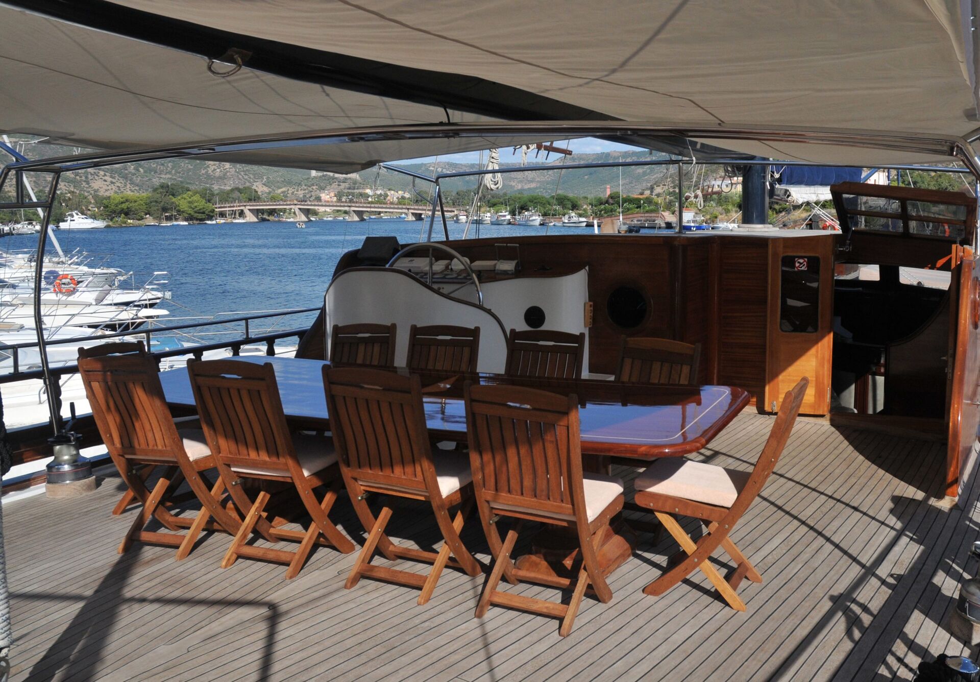 A large wooden dining table with ten chairs is set on the deck of a yacht, overlooking calm blue water and a distant shoreline under a sunshade.