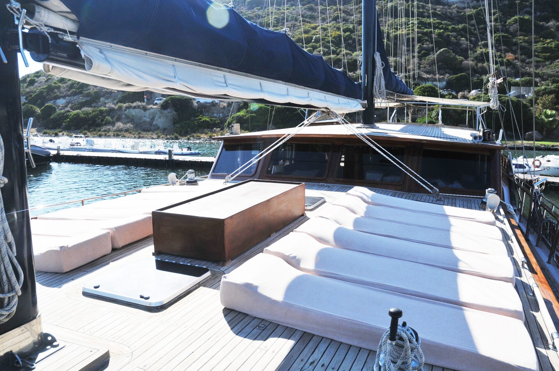 A view from the deck of a yacht featuring cushioned lounging areas and a wooden table, with water and greenery-covered hills visible in the background.