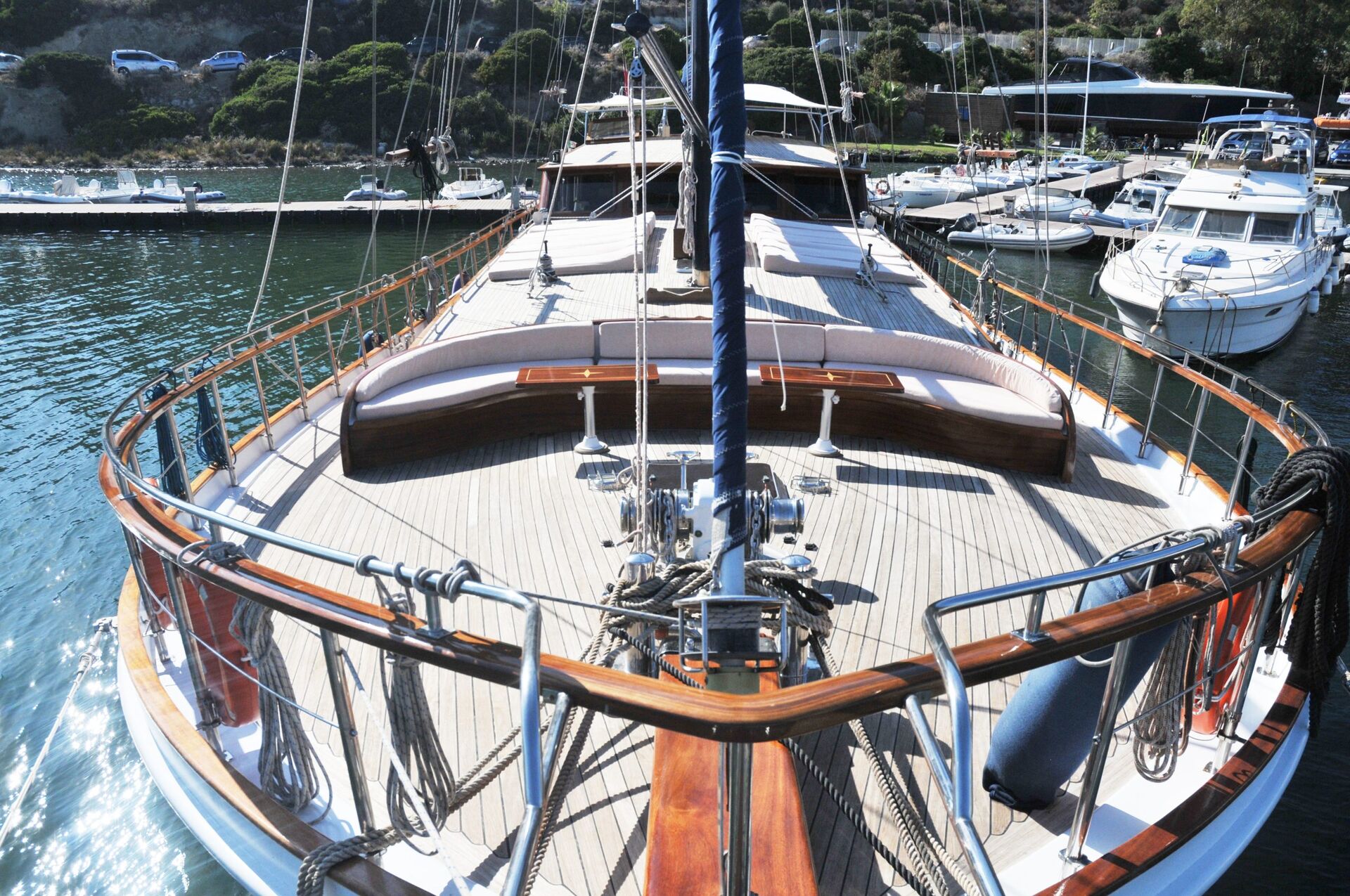 View from the bow of a wooden-decked sailboat docked in a marina, surrounded by several other boats and clear, greenish-blue water under a sunny sky.