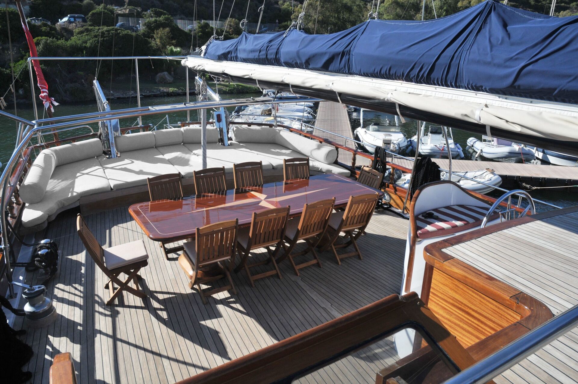 A large wooden dining table with multiple chairs is set on the deck of a yacht, with cushioned seating along the sides and covered sails overhead. The yacht is docked near water with other boats nearby.