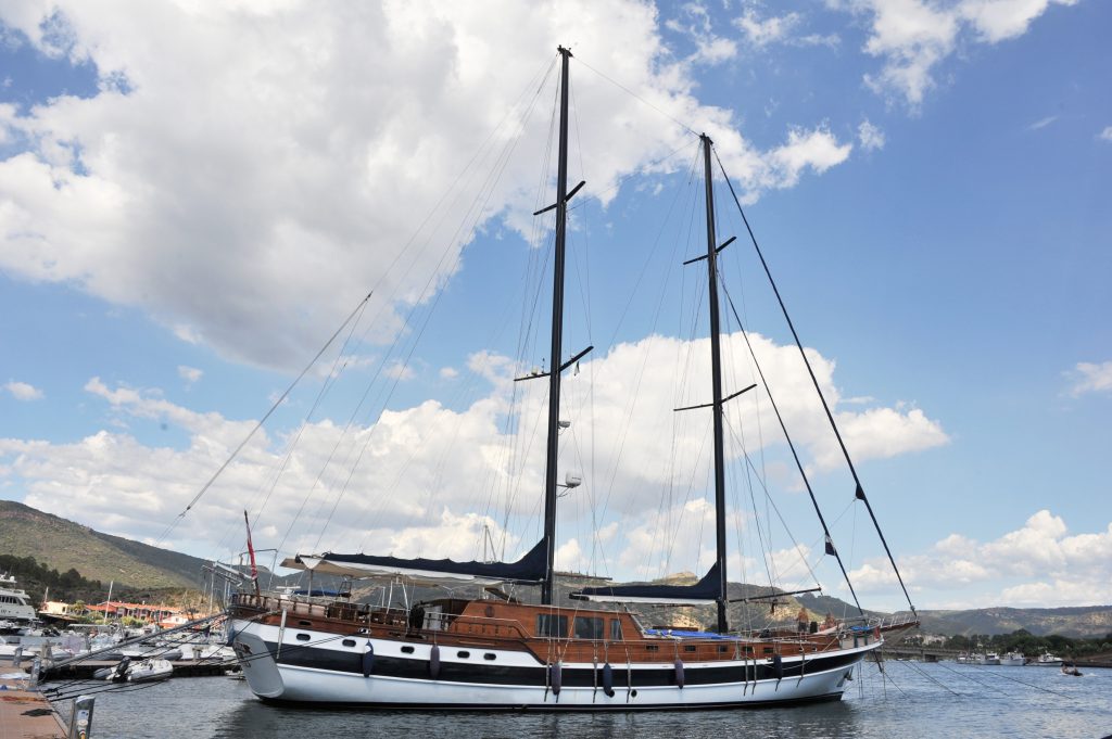 A large sailboat with two tall masts is docked at a marina under a partly cloudy sky, with hills and buildings visible in the background.