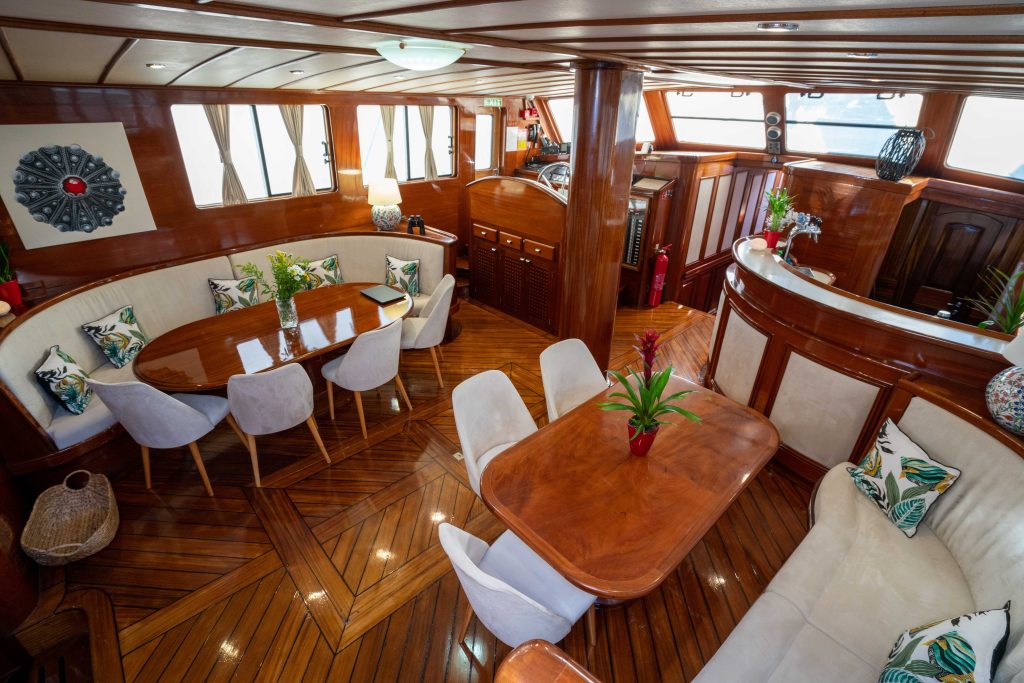 A luxurious yacht interior with polished wood floors, a curved dining area with white chairs, a cozy seating nook, tropical decor, and large windows letting in natural light.