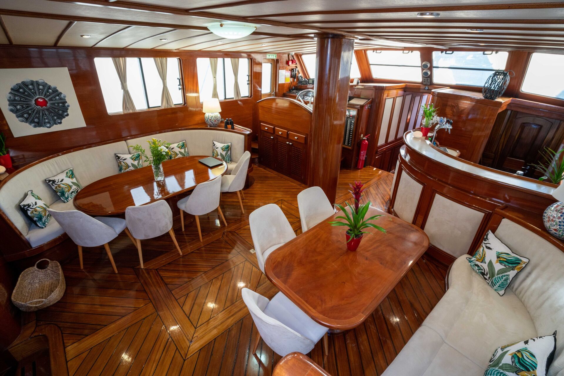 A luxurious yacht interior with polished wood floors, a curved dining area with white chairs, a cozy seating nook, tropical decor, and large windows letting in natural light.