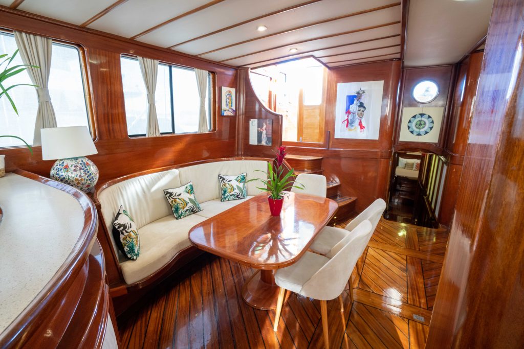 Luxurious yacht interior with glossy wood paneling, a curved white sofa with decorative pillows, a wooden dining table, two white chairs, and large windows letting in natural light. Art and decor accent the walls.