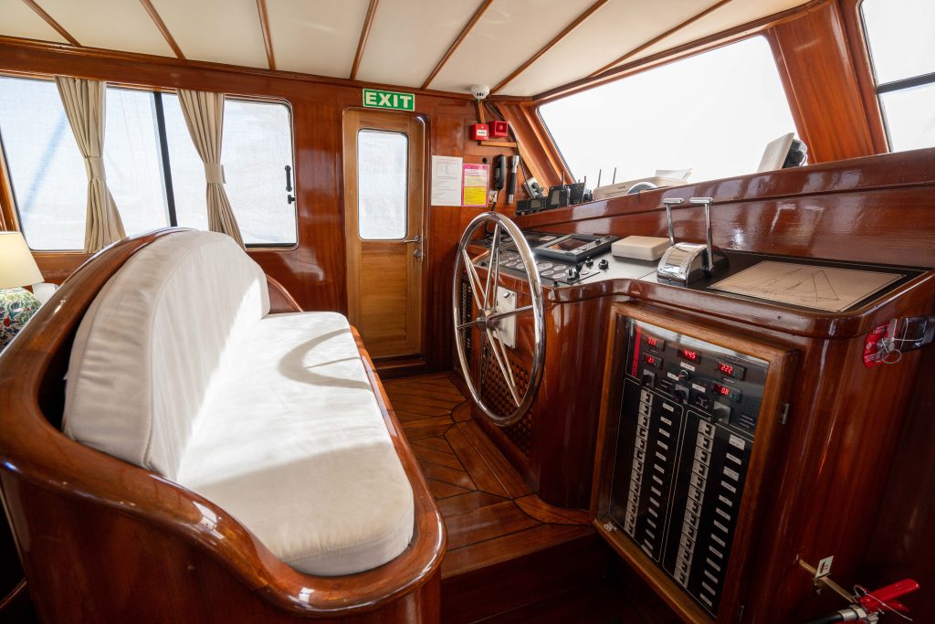The interior of a boat’s helm with a cushioned wooden bench, a large steering wheel, navigation instruments, control panels, and a door with an exit sign above it. Sunlight filters through curtained windows.