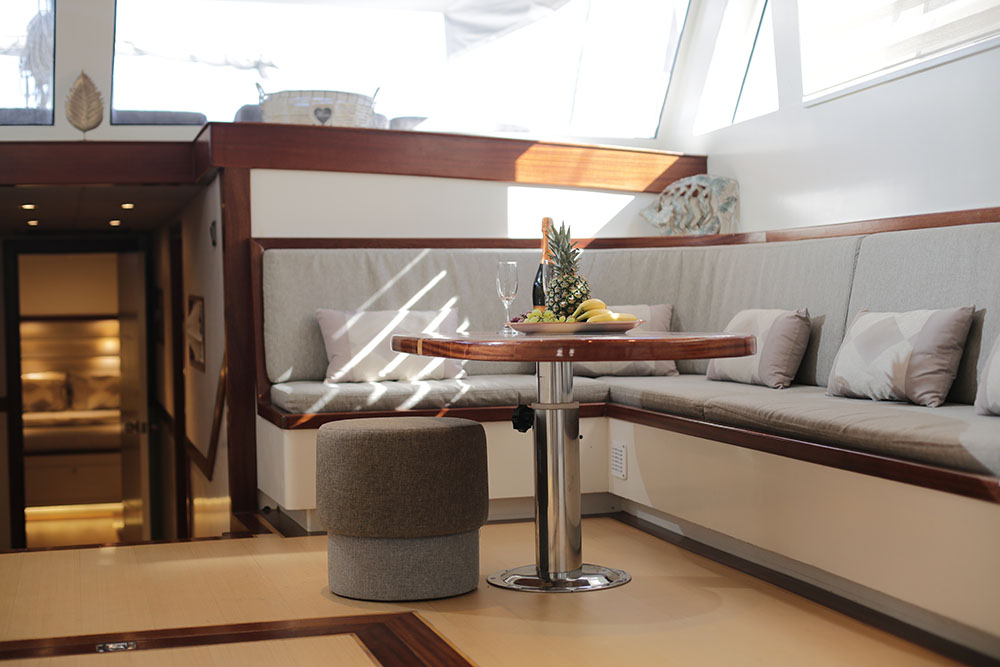A modern yacht interior with a cushioned seating area, a round wooden table set with fruit and a wine glass, soft gray pillows, and bright natural light from large windows.