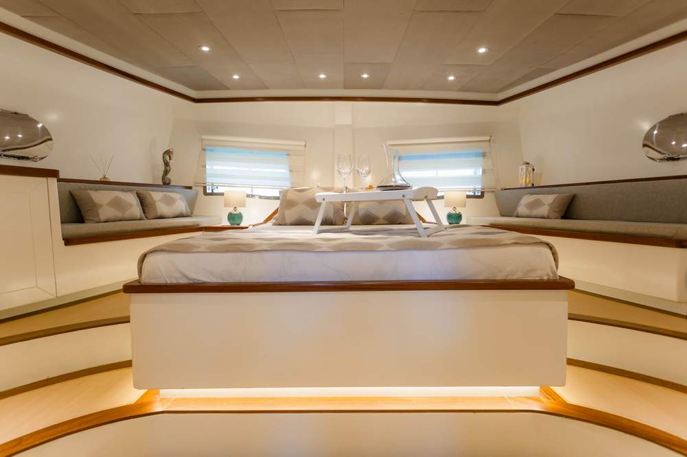 A luxurious bedroom on a yacht features a large bed with a breakfast tray and two wine glasses, built-in seating with cushions along the sides, soft lighting, and two windows with blinds.