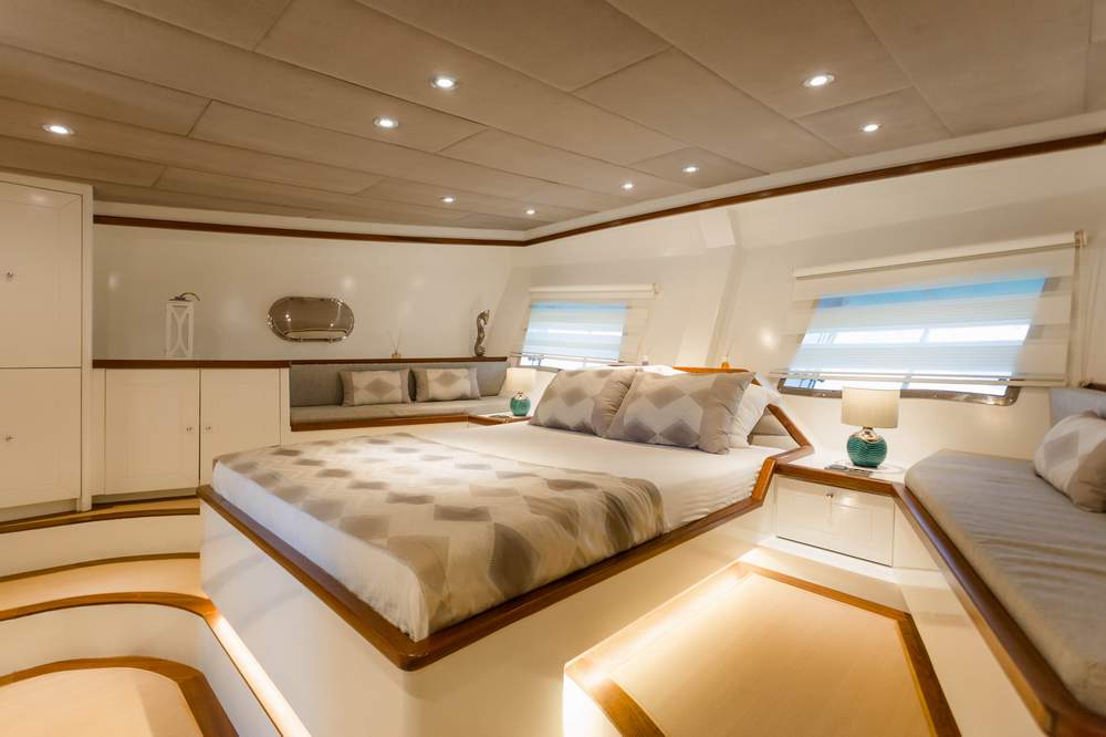 A luxurious, modern yacht bedroom with a large bed, soft lighting, two windows with blinds, built-in cabinets, a cushioned seating area, and stylish decor in neutral tones.