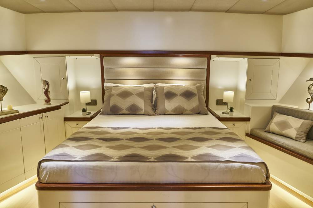 A modern, neatly-made bed with patterned bedding is centered in a cozy bedroom. There are two matching pillows, two bedside tables with lamps, and built-in cabinets. The lighting is warm and the decor is minimal.