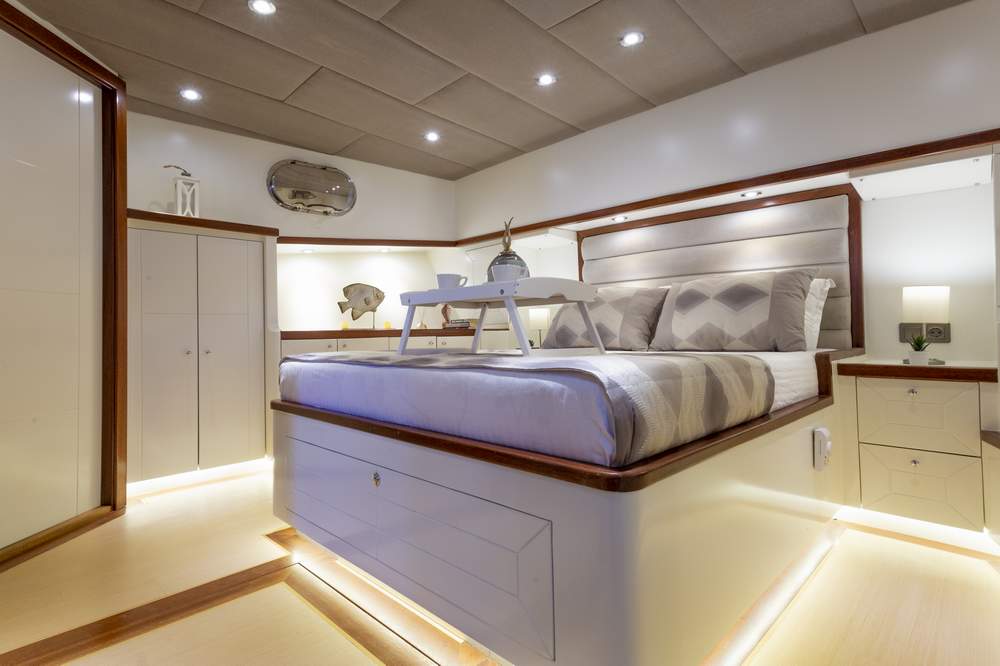 A modern, well-lit bedroom with a raised platform bed, neutral bedding, a breakfast tray, and built-in storage cabinets. Warm recessed lighting highlights the clean, minimalist decor.