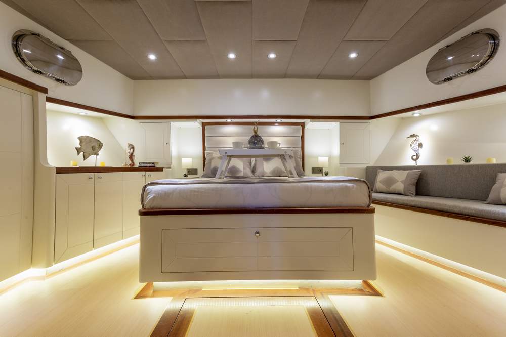 Luxurious yacht bedroom with a large bed, soft lighting, beige and white decor, cushioned bench seating, nautical-themed art, and porthole windows on both sides of the room.