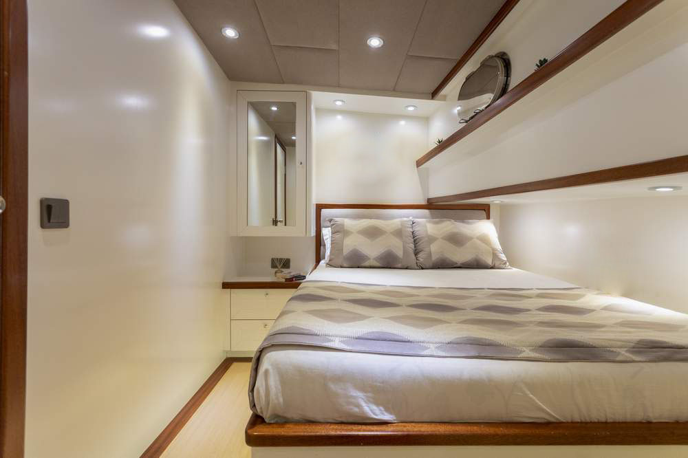 Modern, minimalistic bedroom with a double bed, geometric-patterned bedding, floating wooden shelves, recessed lighting, a small nightstand, and a mirror on the wall, all in a neutral color palette.