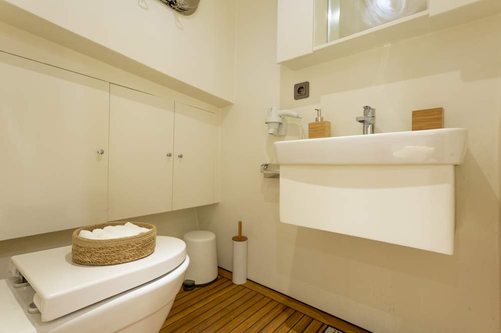 A modern, minimalist bathroom with a white toilet, wicker basket, white sink, wooden soap dispensers, and a wooden slat floor. White cabinets and a wall mirror are visible above the sink.