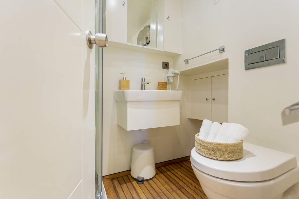 Modern, compact bathroom with a white sink, mirror, and storage cabinets. Toilet with a basket of rolled white towels on top, and wooden flooring. Soap dispensers and a towel bar are near the sink.