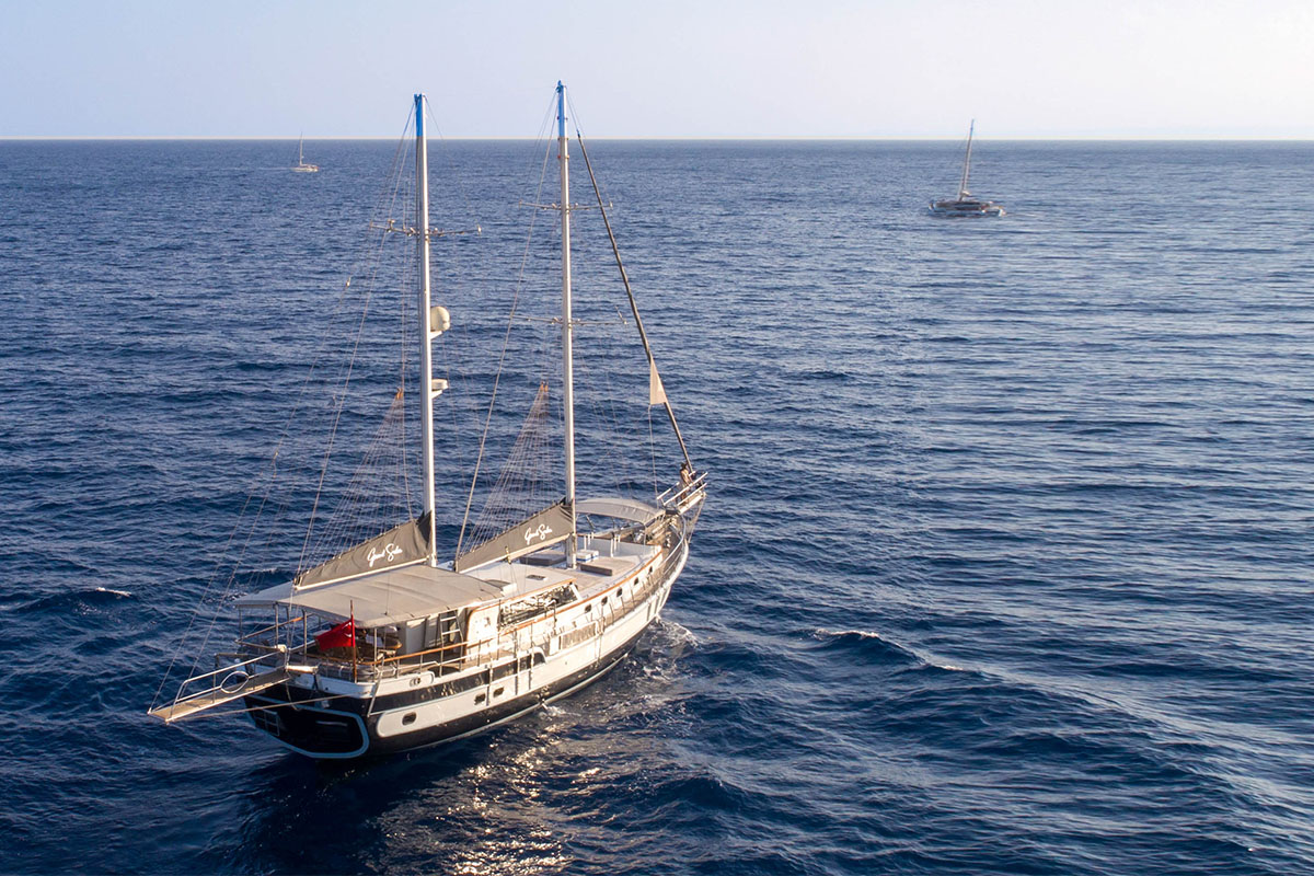 A large sailboat with two masts floats on calm blue water, with two other sailboats visible in the distance under a clear sky.