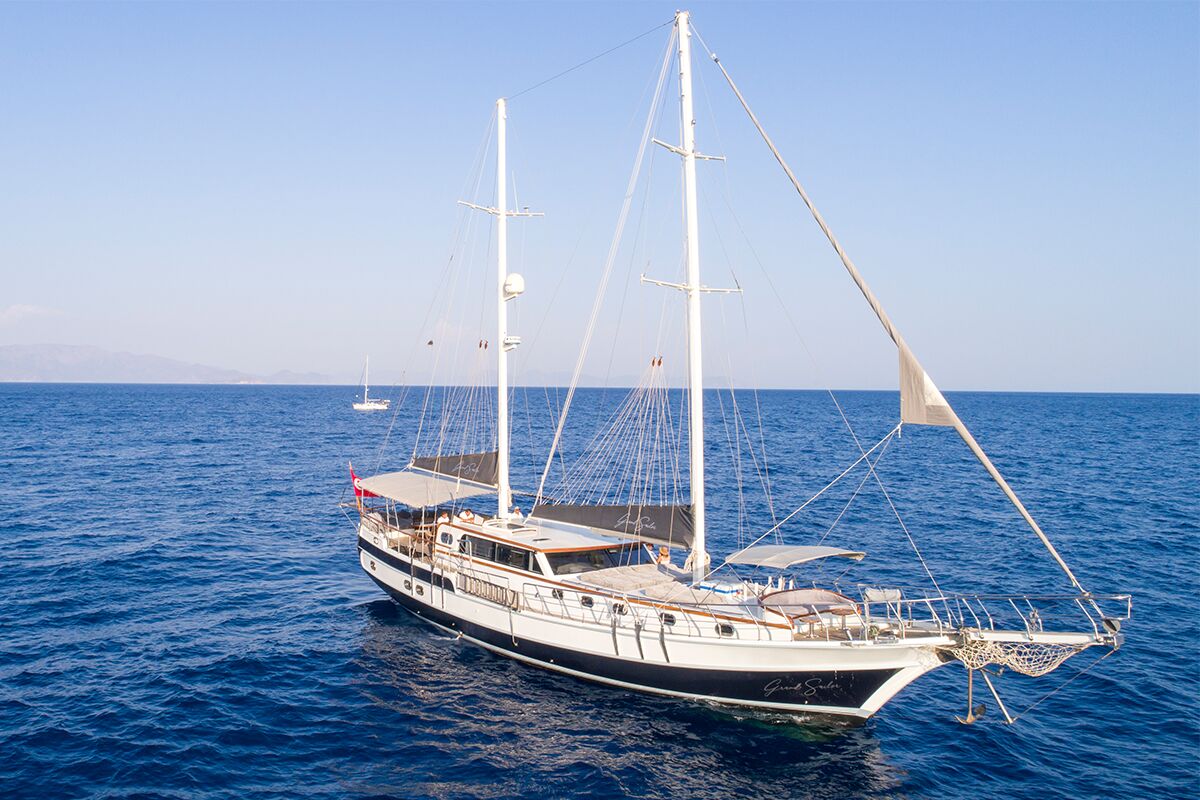 A large sailboat with two masts floats on calm blue sea under a clear sky. The yacht has a sleek black and white hull and various shaded deck areas. Mountains are faintly visible on the distant horizon.