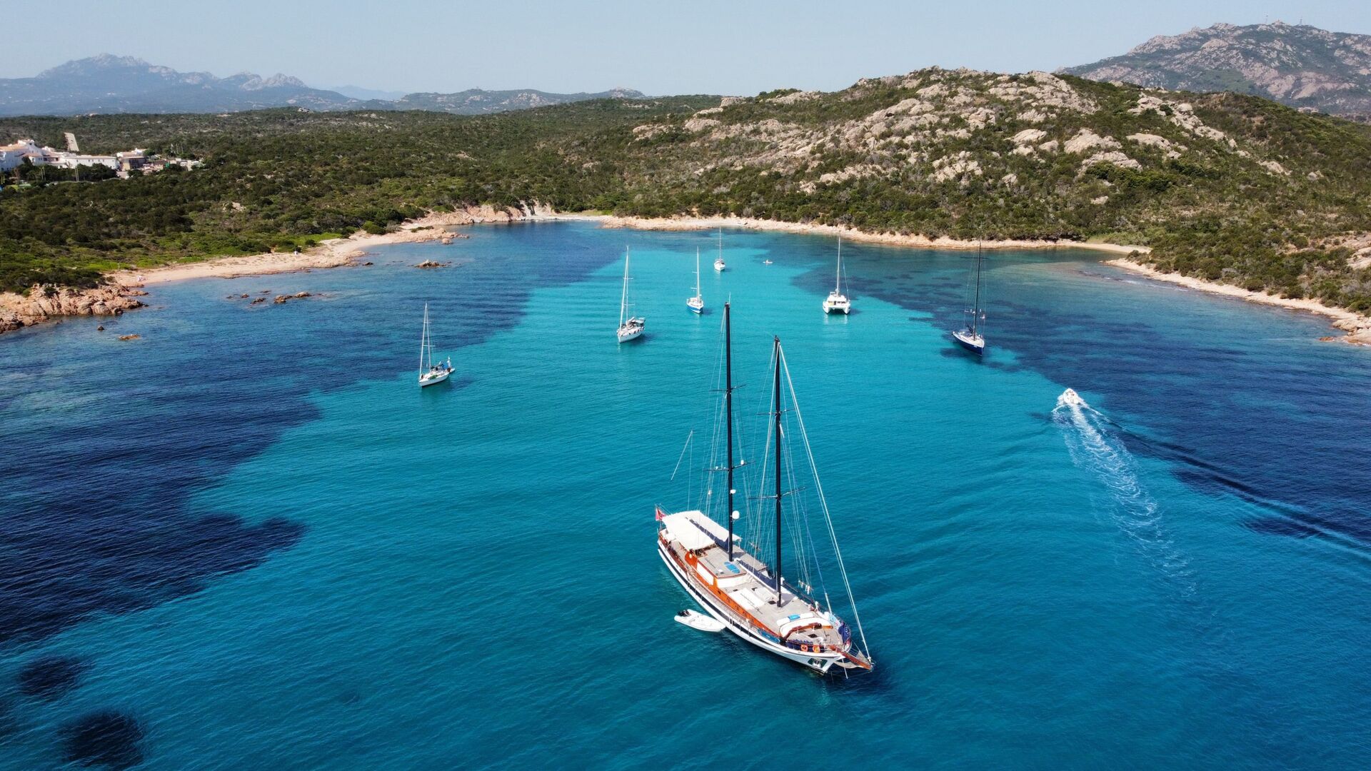 Aerial view of a sailboat and several smaller boats anchored in a clear, turquoise bay surrounded by green hills and rocky coastline under a blue sky.