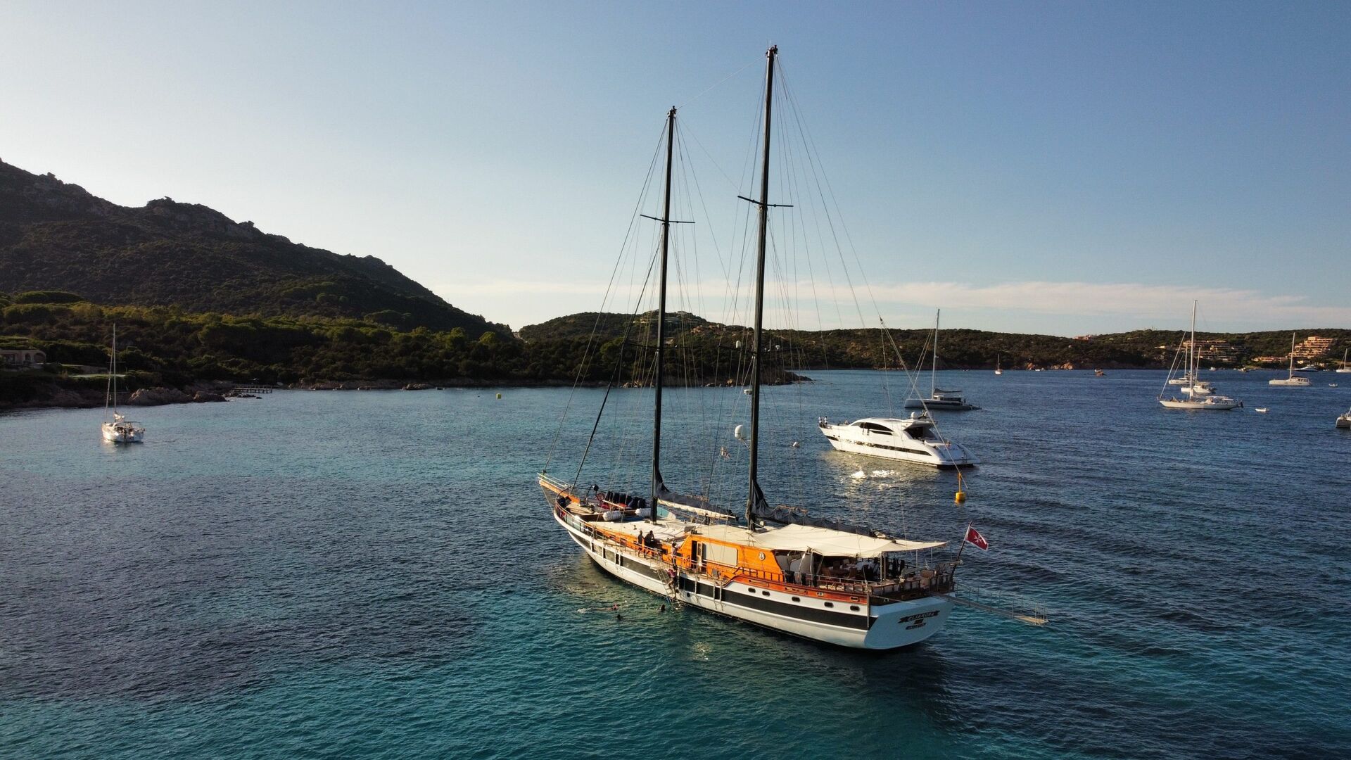 A large sailboat with two masts is anchored in clear blue water near a coastline with hills and trees. Several other boats and yachts are in the background under a clear sky.