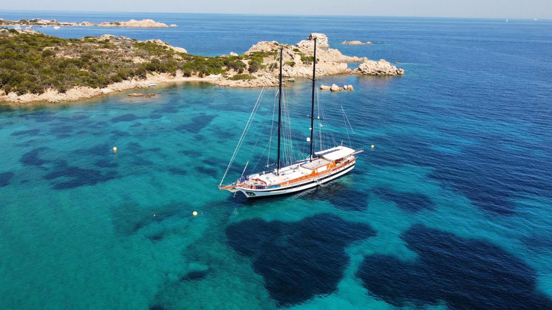 A large sailboat floats on clear turquoise water near a rocky, green island, with shallow areas and deeper blue patches visible in the sea under a sunny sky.