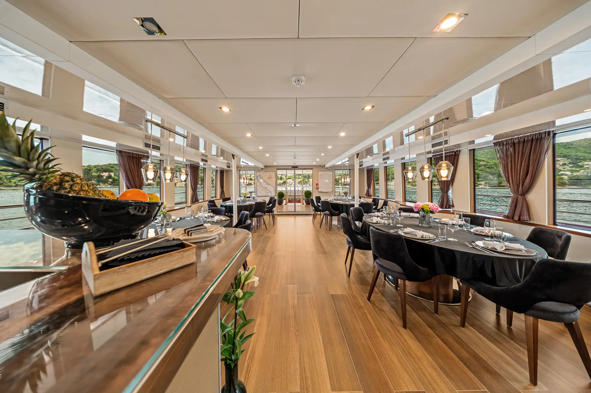Elegant dining area on a yacht with black chairs, set tables, large windows showing water and hills outside, wooden flooring, soft lighting, and decorative fruit bowls and flowers.