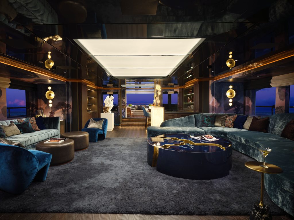 A luxurious yacht lounge with plush blue and gray sofas, modern gold and glass accents, soft lighting, and large windows displaying an ocean view at dusk. A sculpted statue and coffee tables complete the elegant setting.