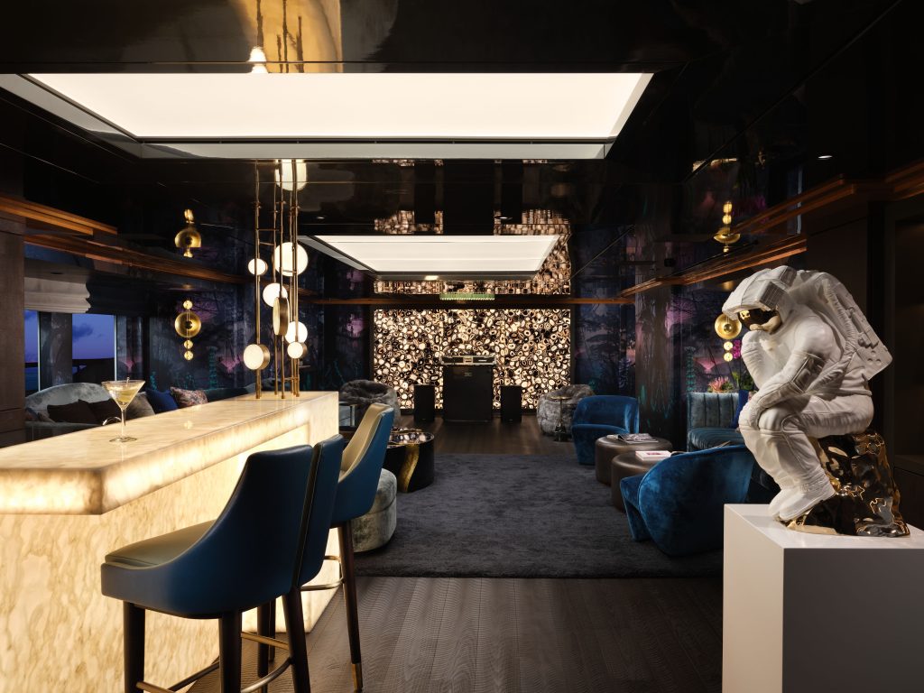 A modern lounge with a glowing marble bar, two blue bar stools, plush seating, a dark rug, and a white astronaut sculpture in a thinking pose. The back wall is illuminated with decorative lights.