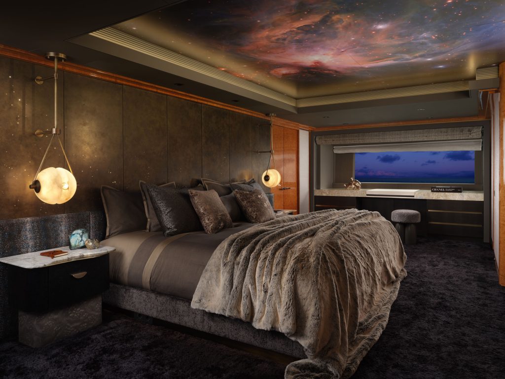 Luxurious bedroom with a fur throw on the bed, plush pillows, two round pendant lights, dark carpeting, and a ceiling featuring a vivid space nebula mural. A large window offers a view of the evening sky.