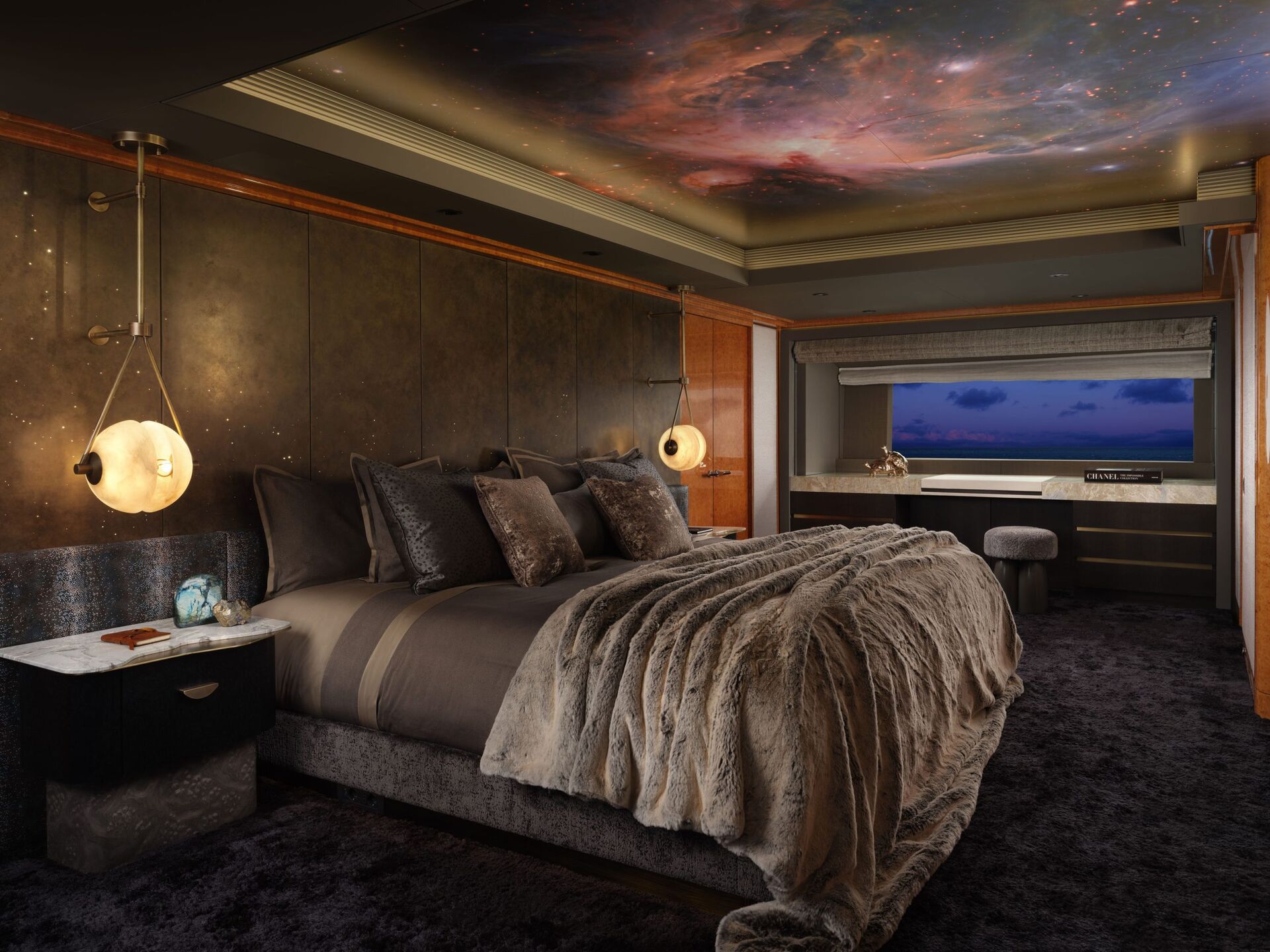 Luxurious bedroom with a fur throw on the bed, plush pillows, two round pendant lights, dark carpeting, and a ceiling featuring a vivid space nebula mural. A large window offers a view of the evening sky.