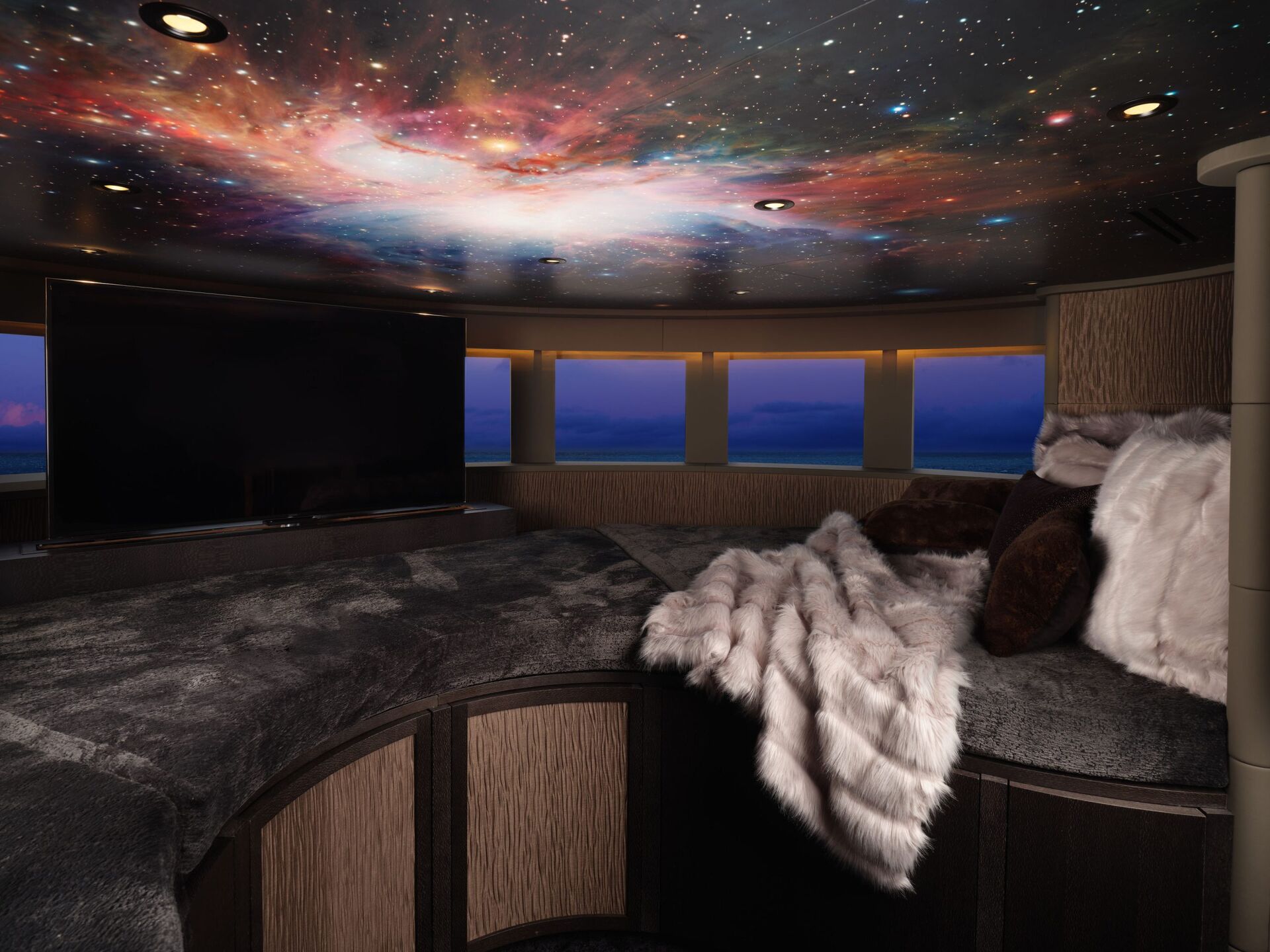 A cozy, modern lounge with a plush fur blanket and pillows, large TV, panoramic windows showing a twilight sky, and a ceiling painted with a vibrant galaxy and stars.