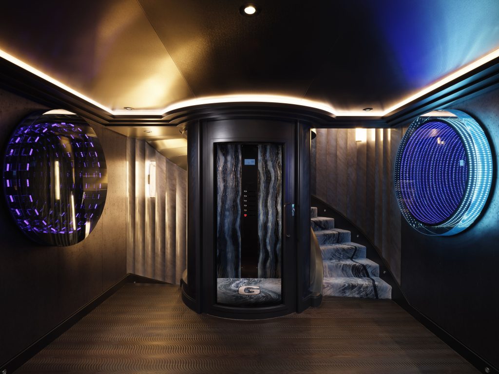 A luxurious hallway features a sleek glass elevator in the center, flanked by blue and purple circular LED light displays on textured walls, with a curved staircase to the right and modern ceiling lighting above.