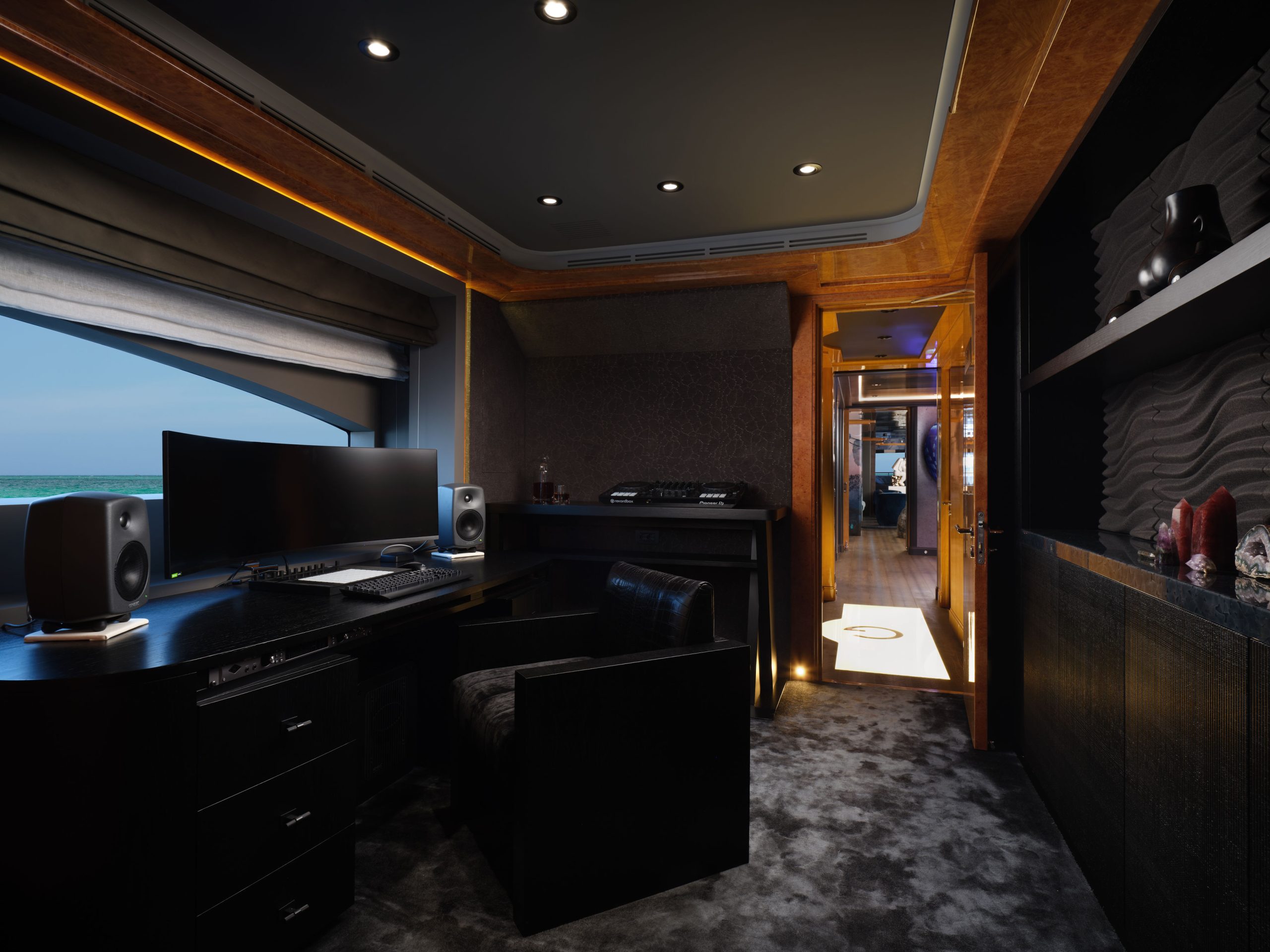 A modern, dimly lit home office with a large desk, dual monitors, speakers, and a black chair. A window shows an ocean view. The room has dark walls, plush carpet, and leads to a well-lit hallway.