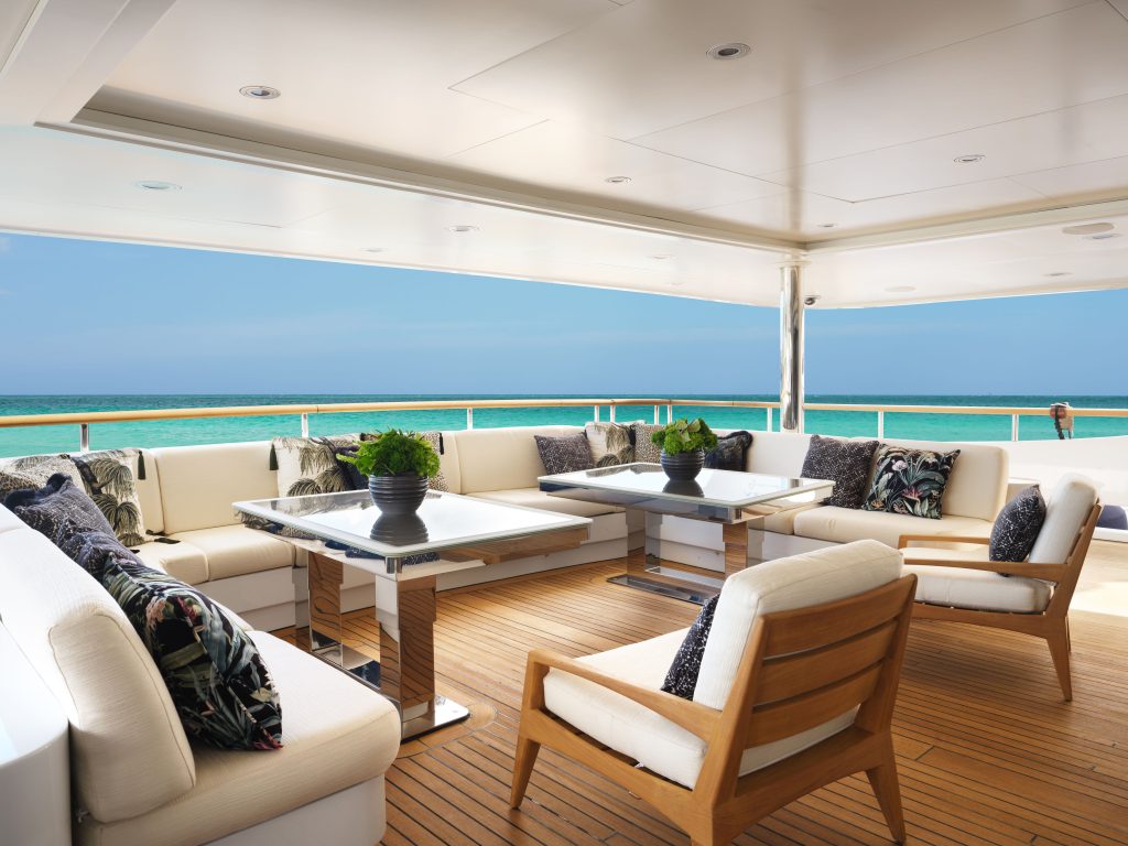 Spacious yacht deck with cushioned seating, wooden chairs, glass tables, and potted plants, overlooking a turquoise ocean under a bright blue sky.