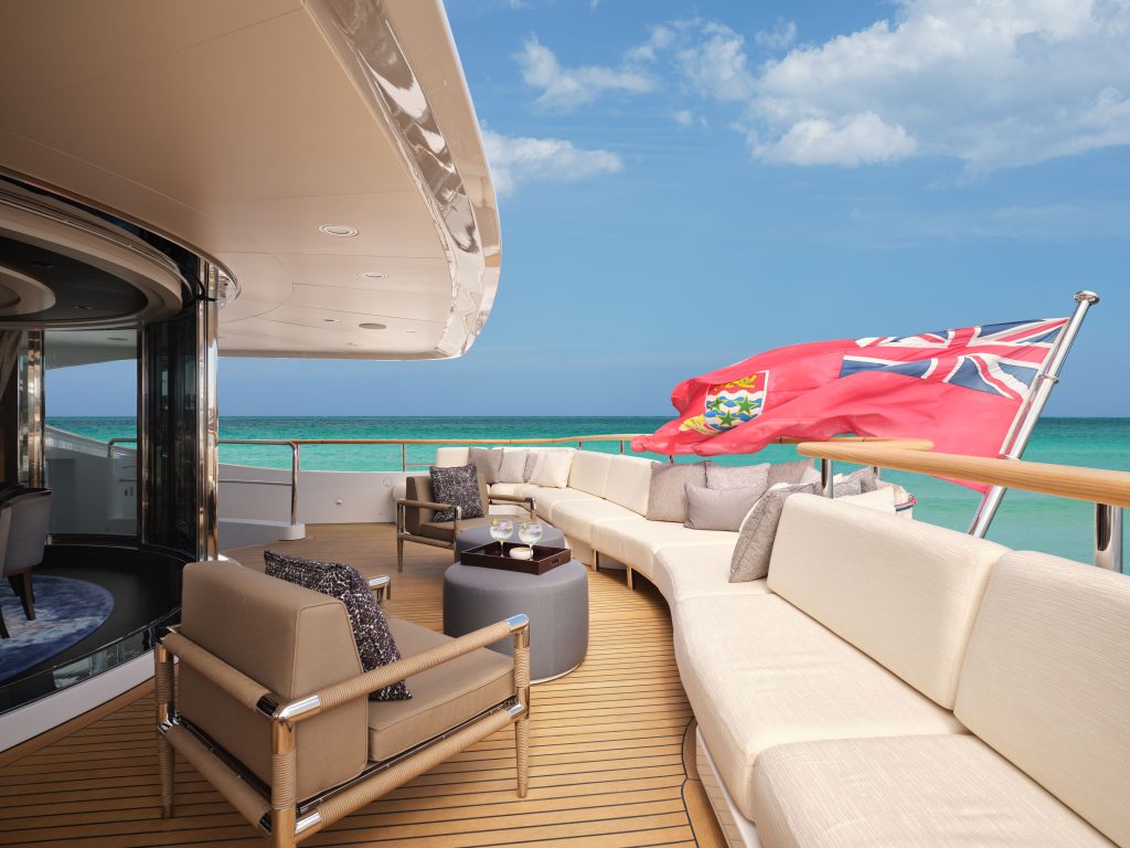Luxurious yacht deck with cream-colored seating, small tables with drinks, and a British Red Ensign flag. The yacht overlooks turquoise ocean water under a sunny, partly cloudy sky.