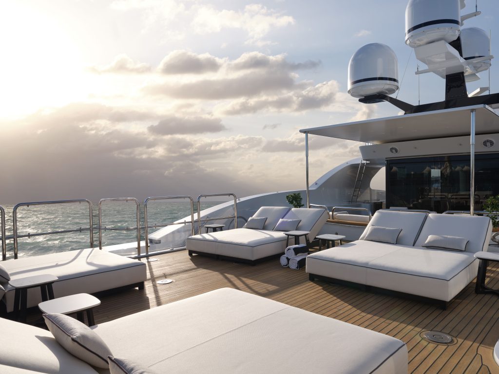 A luxurious yacht deck with large white lounge beds, modern railings, and satellite domes, overlooking a calm ocean at sunset under a partly cloudy sky.