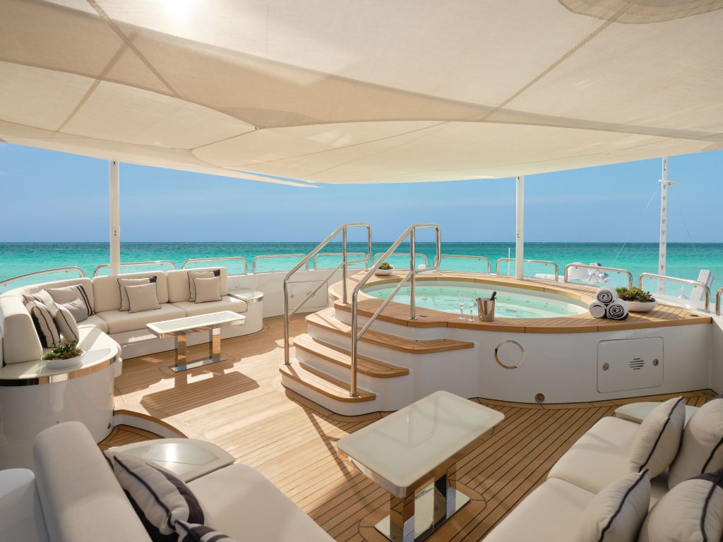 A luxurious yacht deck with plush seating, tables, and a round hot tub in the center, overlooking clear turquoise ocean water under a white canopy on a sunny day.
