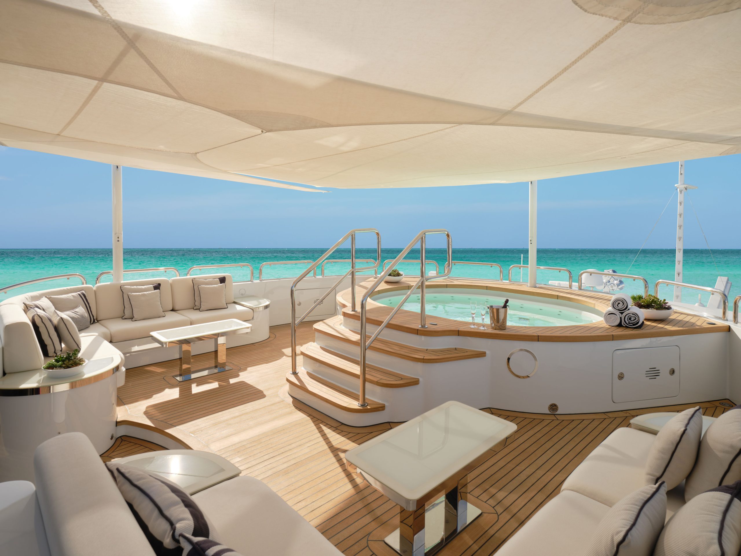 A luxurious yacht deck with plush seating, tables, and a round hot tub in the center, overlooking clear turquoise ocean water under a white canopy on a sunny day.