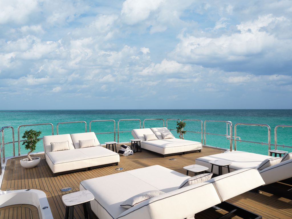 Spacious yacht deck with cushioned lounge chairs and tables, overlooking clear turquoise ocean water under a partly cloudy sky. Two potted plants add a touch of greenery.