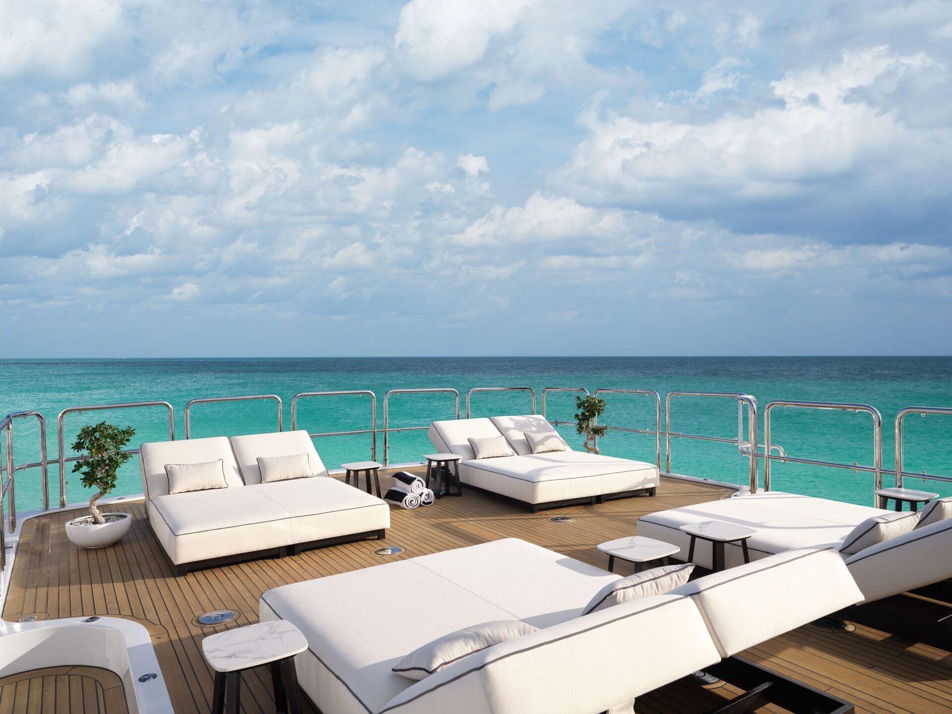 Spacious yacht deck with cushioned lounge chairs and tables, overlooking clear turquoise ocean water under a partly cloudy sky. Two potted plants add a touch of greenery.