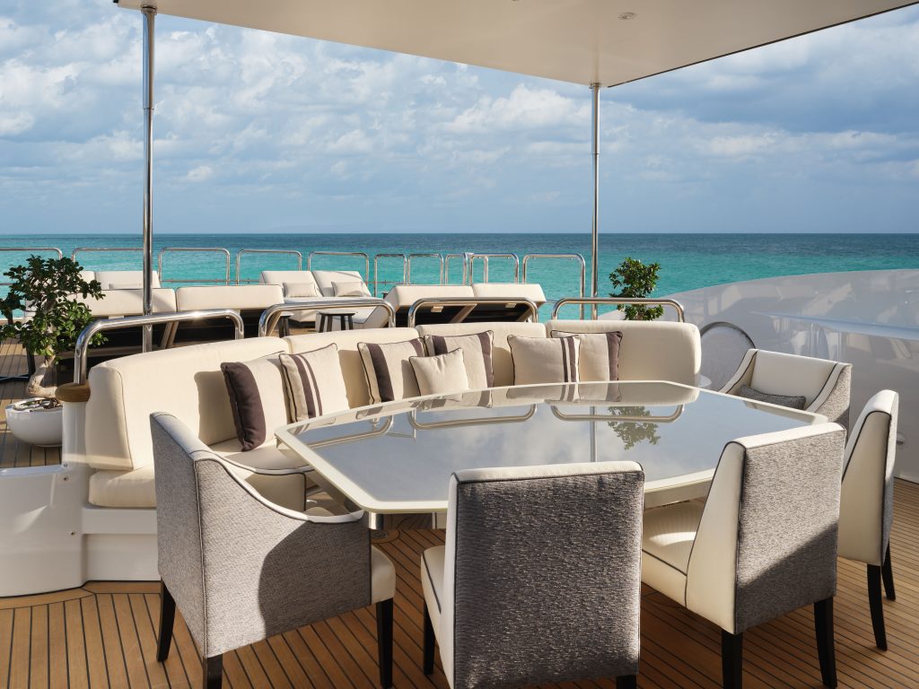 A luxurious outdoor yacht dining area with cushioned chairs and sofas around a glossy table, overlooking turquoise ocean water under a partly cloudy sky. Potted plants add a touch of greenery to the elegant setting.