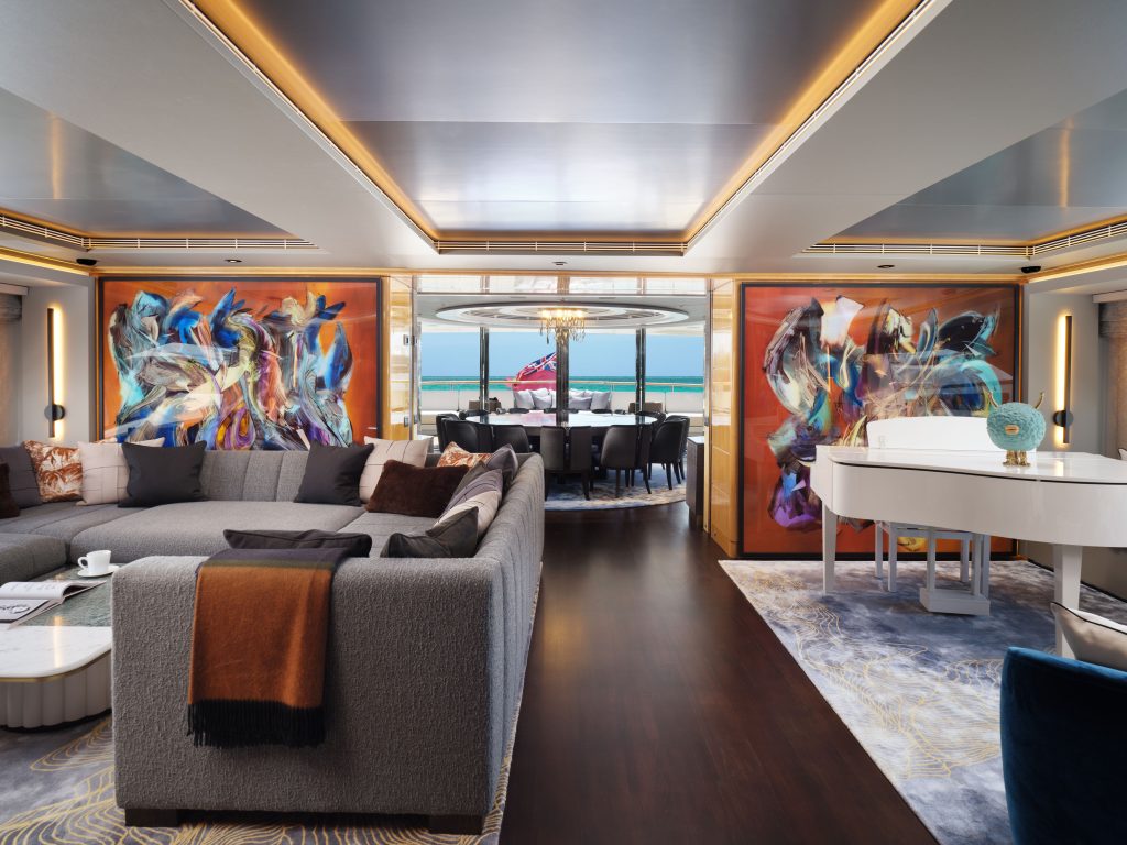 A luxurious yacht interior with a spacious living area, modern gray sectional sofa, white grand piano, abstract wall art, and large windows offering a sea view. A dining area is visible in the background with ocean beyond.