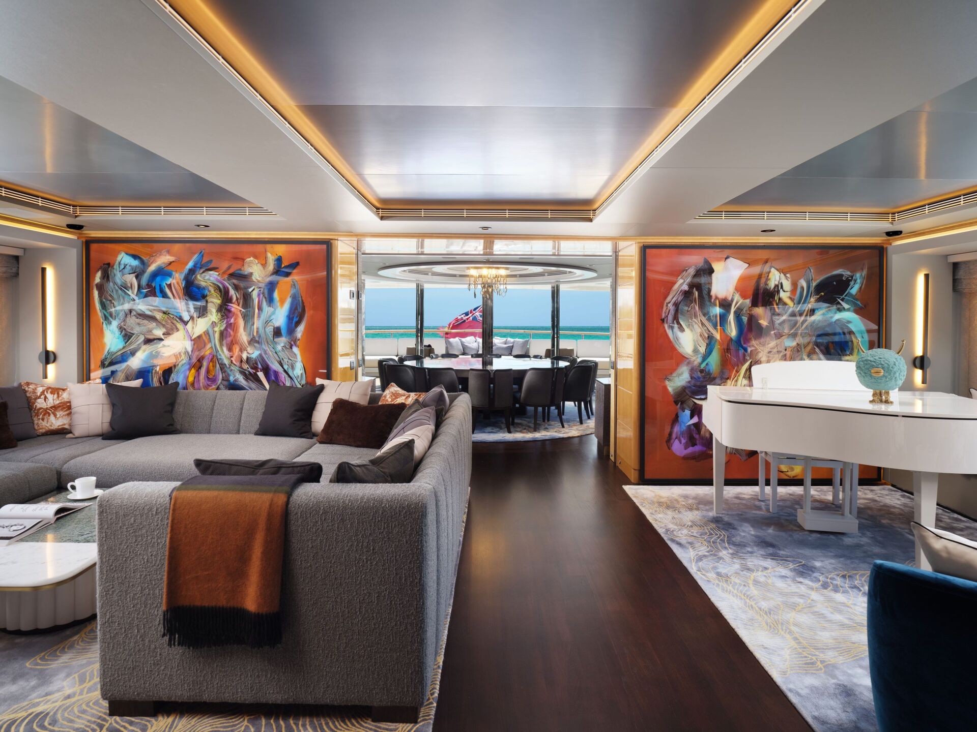 A luxurious yacht interior with a spacious living area, modern gray sectional sofa, white grand piano, abstract wall art, and large windows offering a sea view. A dining area is visible in the background with ocean beyond.