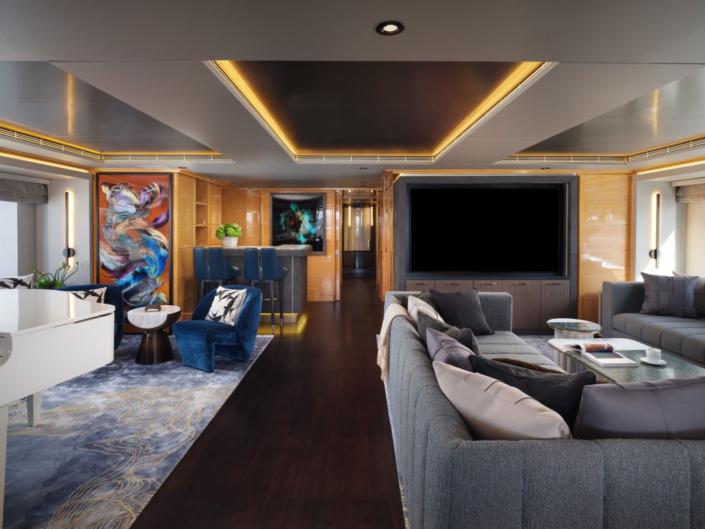 Modern, luxurious living room with dark wood floors, gray sofas, a white piano, blue accent chairs, abstract wall art, a large flat-screen TV, bar area, and warm recessed lighting.