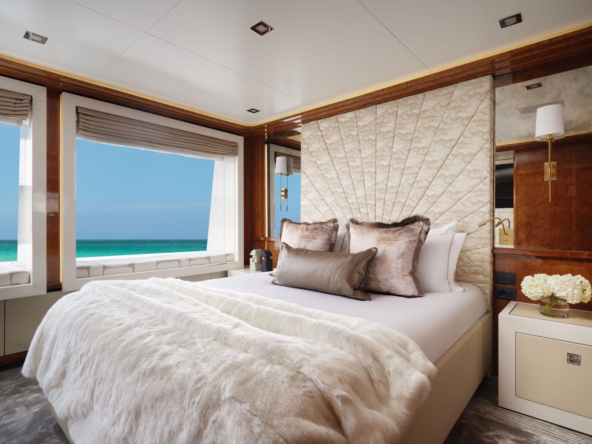 Luxurious yacht bedroom with a plush bed, soft bedding, pillows, and elegant decor. Large windows offer a stunning view of the turquoise ocean and blue sky, filling the room with natural light.