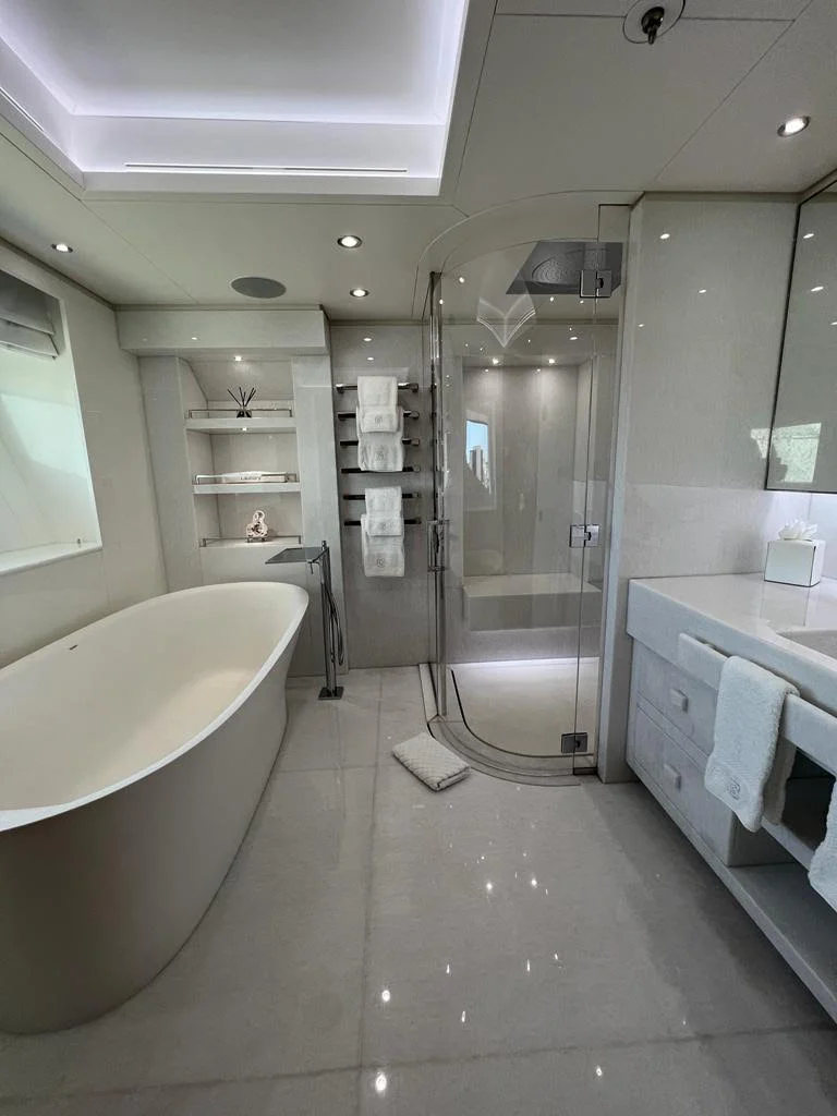 Modern bathroom with a freestanding bathtub, glass shower, wall-mounted towel rack, shelves with decor items, vanity with sink, and white towels, all in a neutral color palette with bright lighting.
