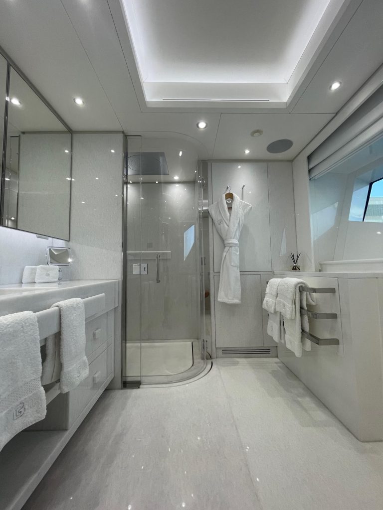 A modern, luxurious bathroom with white and light gray decor, featuring a glass-enclosed shower, double sinks with towels, a bathrobe hanging on the wall, and a large window allowing natural light inside.