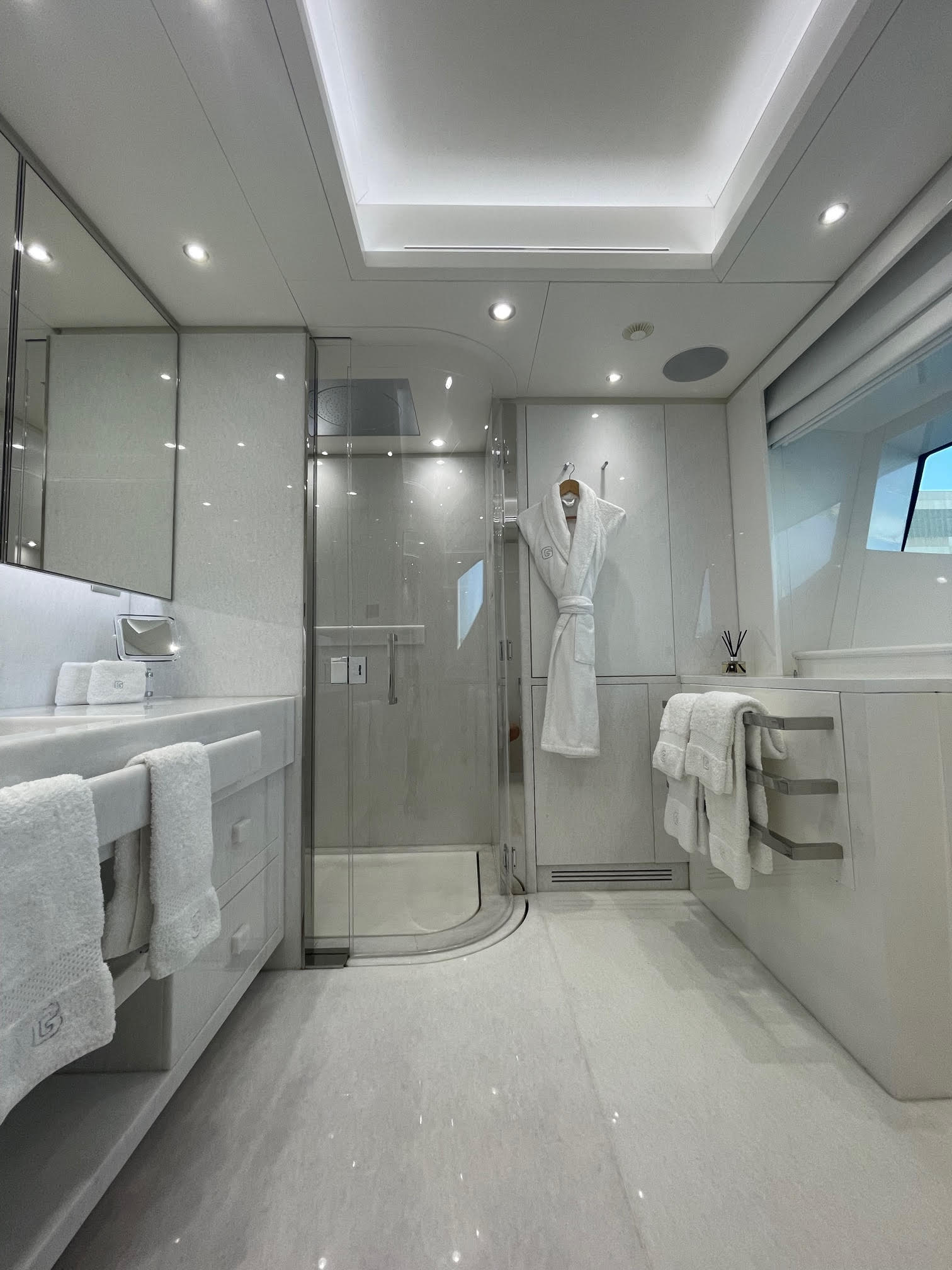 A modern, luxurious bathroom with white and light gray decor, featuring a glass-enclosed shower, double sinks with towels, a bathrobe hanging on the wall, and a large window allowing natural light inside.